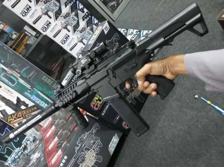 Pistola de hidrogel Arp9 negra con mira holográfica y accesorios tácticos, sostenida por una mano sobre cajas de empaque.