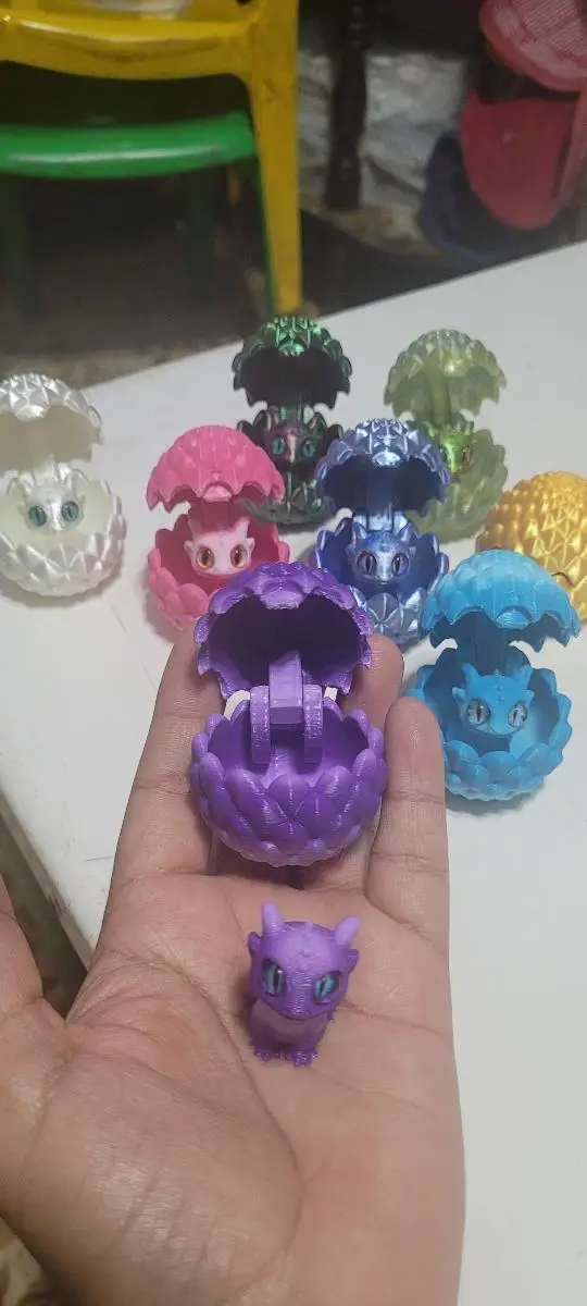 Mano sosteniendo huevo dragón mini 3D morado abierto con su pequeña figura, rodeado de otros huevos de colores con textura de escamas.