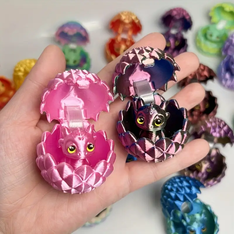 Mano sosteniendo huevos dragón mini 3D abiertos en colores rosa y morado metálico, con pequeños dragones interiores y otras variantes al fondo.