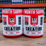 Set de dos tarros de Creatina Monohidrato Old School con BioFit de 300g, color blanco y rojo, sobre superficie de madera en tienda.