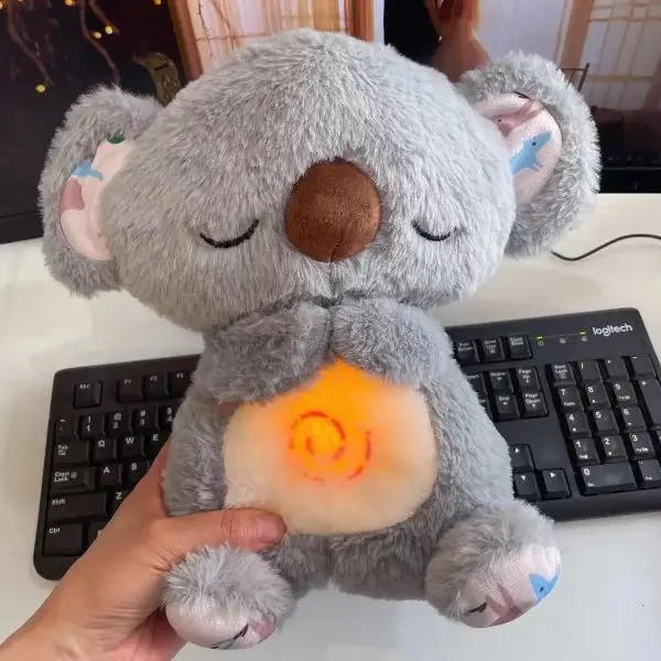 Peluche de koala gris con textura suave, ojos cerrados y luz cálida en espiral sobre el abdomen, sostenido frente a un teclado de computadora.