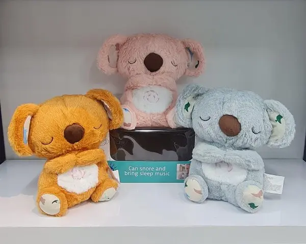 Tres peluches de koala en colores naranja, rosa y gris con función de música y respiración sobre fondo blanco.