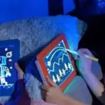 Dos niños dibujan en una pizarra Magic Pad 3D transparente; uno usa el borde azul con dibujo de robot y otro el borde rojo con paisaje iluminado en la oscuridad.