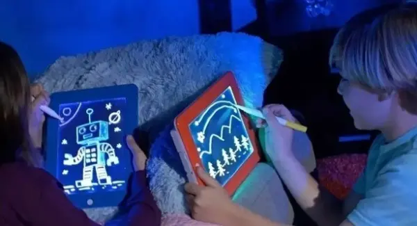 Dos niños dibujan en una pizarra Magic Pad 3D transparente; uno usa el borde azul con dibujo de robot y otro el borde rojo con paisaje iluminado en la oscuridad.