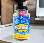 Frasco transparente con tapa negra que contiene cuatro sobres de líquido para burbujas en verde y fucsia, con etiqueta azul y texto "Bubble".