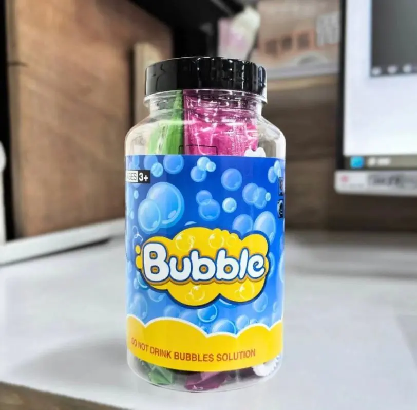 Frasco transparente con tapa negra que contiene cuatro sobres de líquido para burbujas en verde y fucsia, con etiqueta azul y texto "Bubble".