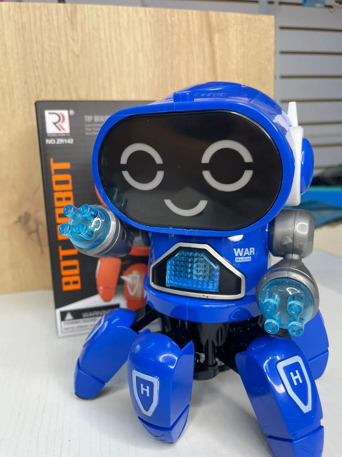 Robot juguete interactivo azul de seis patas con luces LED en las manos y la pantalla frontal, acompañado de su caja de empaque en una superficie blanca.