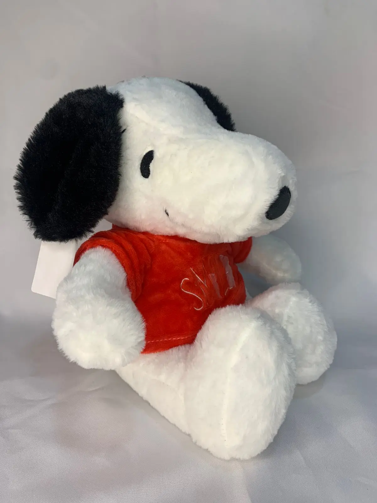 Peluche de Snoopy blanco con orejas negras y camiseta roja afelpada, sentado en perfil, ideal para decoración infantil.