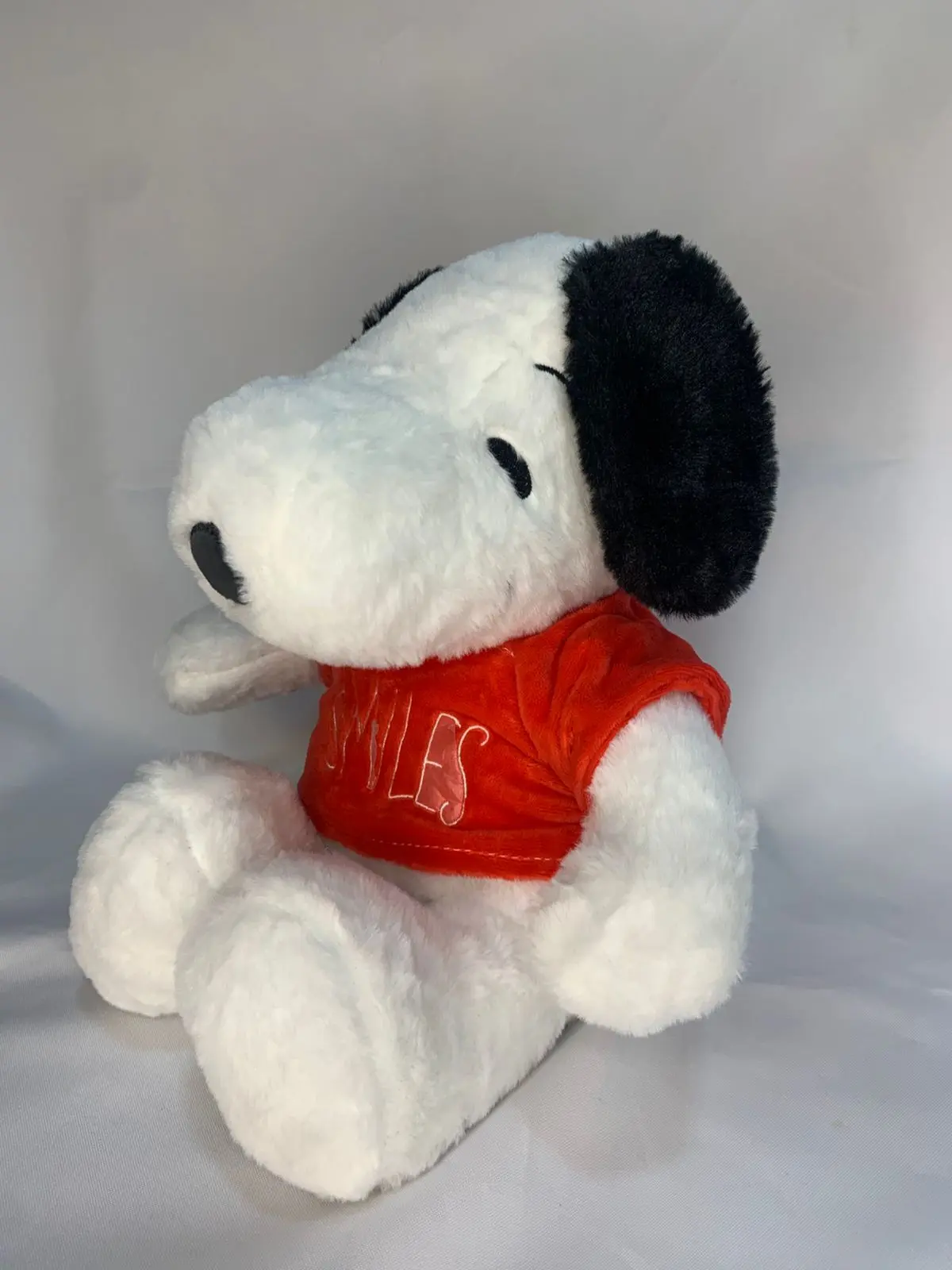 Peluche de Snoopy blanco con orejas negras y camiseta roja afelpada con la palabra SIBILES, en posición sentada sobre fondo blanco.