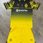 Conjunto de fútbol Borussia Dortmund con camiseta negra y amarilla degradada y shorts amarillos con logos bordados, sobre fondo de madera.