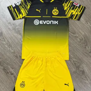 Conjunto de fútbol Borussia Dortmund con camiseta negra y amarilla degradada y shorts amarillos con logos bordados, sobre fondo de madera.
