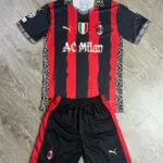 Conjunto de fútbol AC Milan negro y rojo con rayas verticales, cuello y detalles florales blancos, que incluye camiseta y pantaloneta doblados sobre superficie de madera.