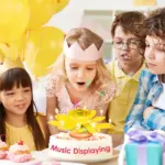 Vela musical de cumpleaños con forma de flor amarilla sobre un pastel blanco, en una fiesta infantil con globos y niños celebrando.