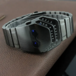 Reloj inteligente de metal gris con diseño futurista hexagonal, pulsera de eslabones y pantalla LED digital sobre fondo de cuero café.