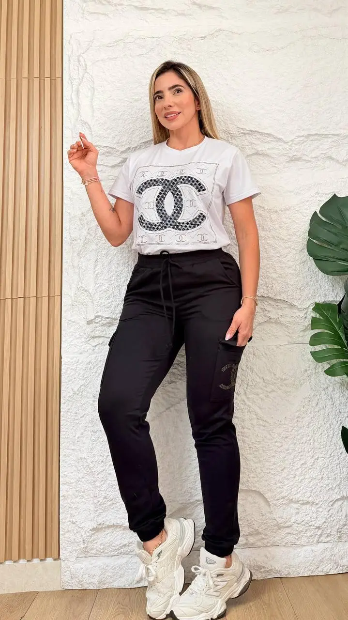 Mujer de pie usando conjunto urbano de lycra negra con jogger cargo y blusón blanco con diseño frontal, zapatillas blancas y fondo texturizado.