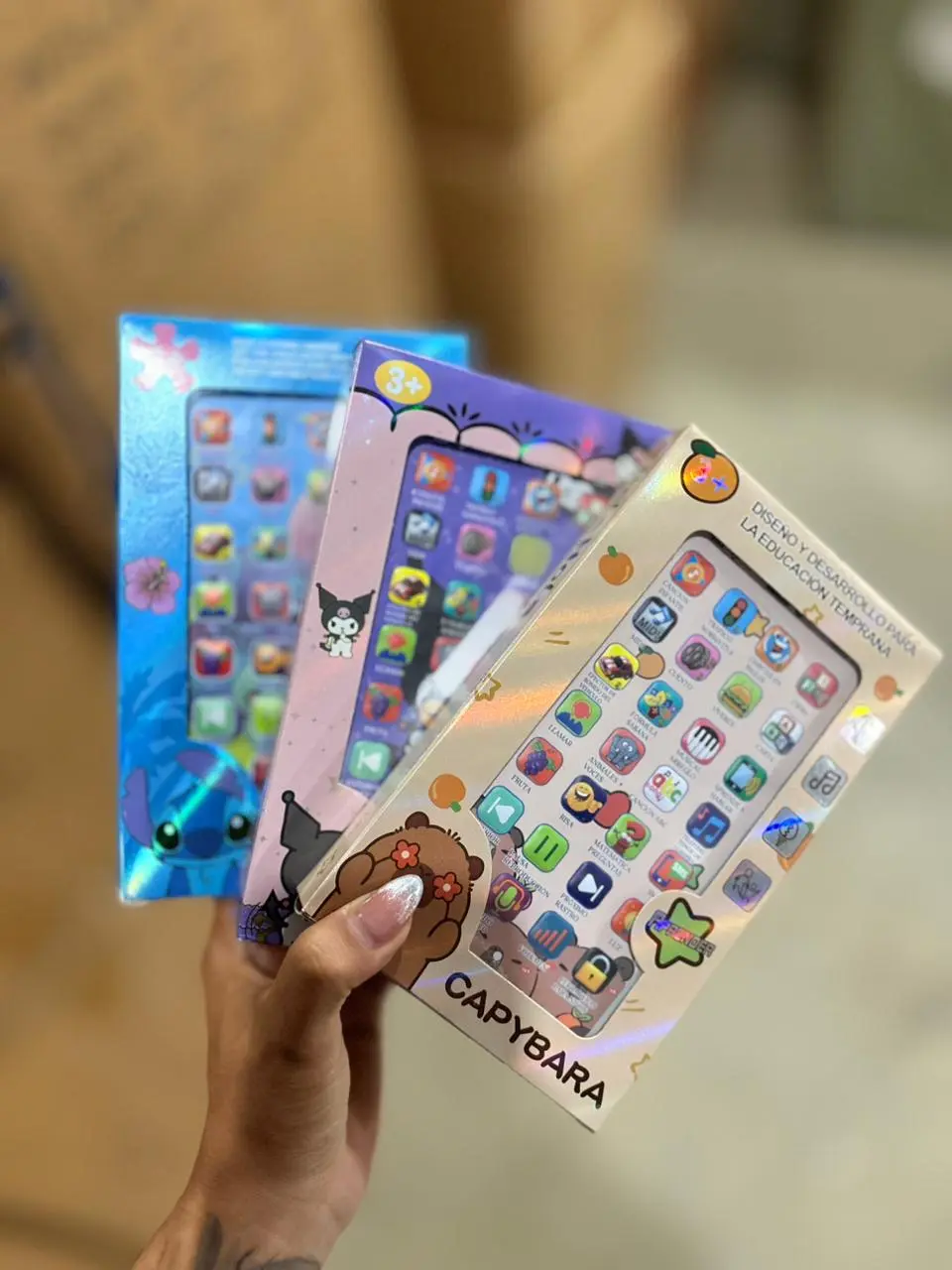Mano sosteniendo tres cajas holográficas de celulares didácticos infantiles con diseños de Stitch, Kuromi y Capybara para aprendizaje temprano.