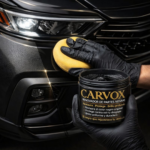 Persona con guantes negros aplicando restaurador Carvox en pasta con una esponja amarilla sobre la parte plástica negra de un carro.
