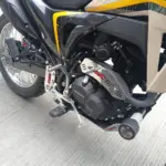 Protector de mofle en acero inoxidable brillante instalado en el escape de una motocicleta, con diseño de ranuras para protección y estética.