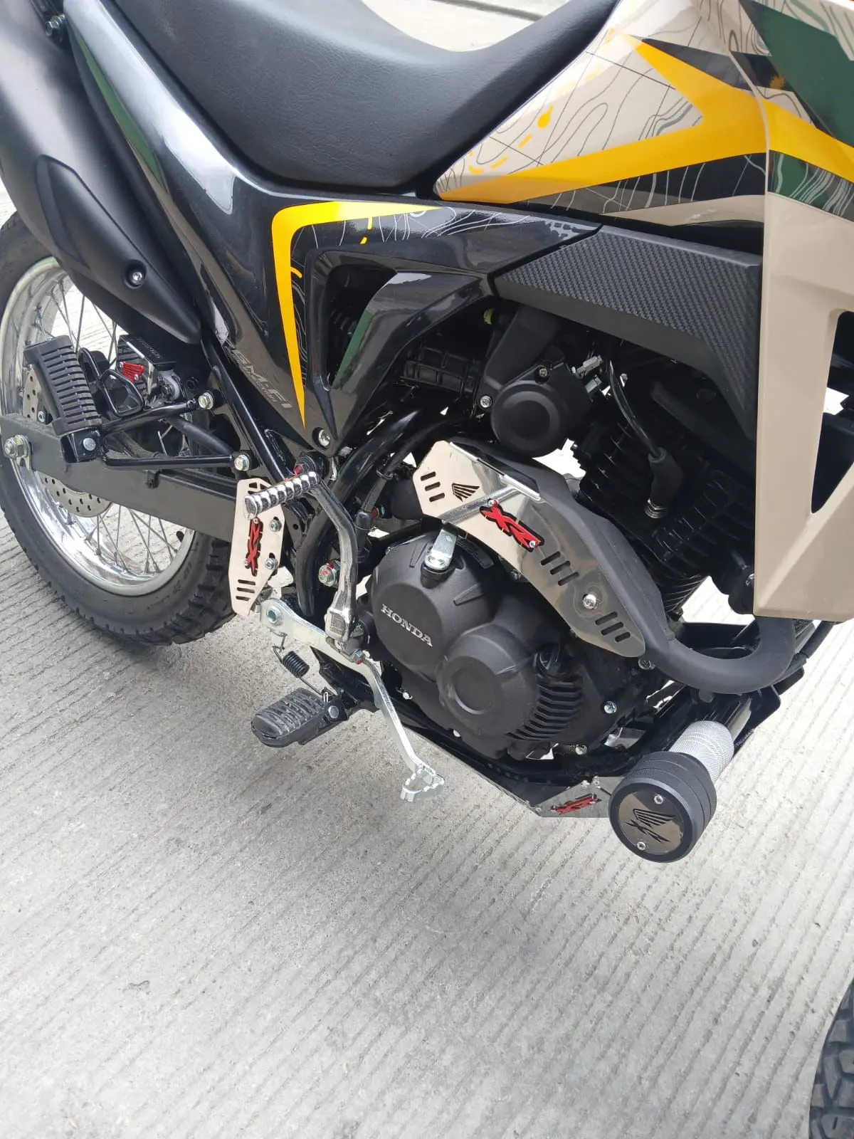 Protector de mofle en acero inoxidable brillante instalado en el escape de una motocicleta, con diseño de ranuras para protección y estética.