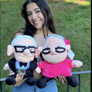 Peluches de Carl y Ellie de Up; uno con traje negro y lentes, otro con vestido rosa y pañoleta, sostenidos por una mujer en un jardín.