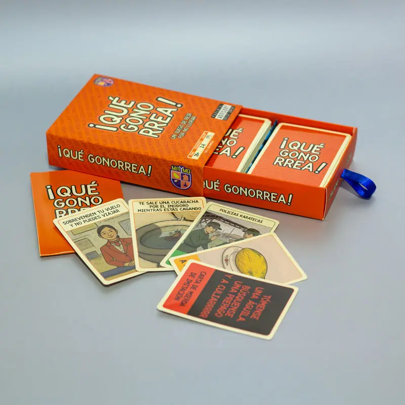 Caja de cartón naranja abierta de juego de mesa ¡Qué Gonorrea!, mostrando cartas ilustradas, manual y una tarjeta negra al frente sobre superficie gris.