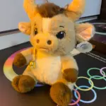 Peluche de toro beige con manchas marrones, cuernos suaves, copete desordenado, campana dorada al cuello y etiqueta colgante.