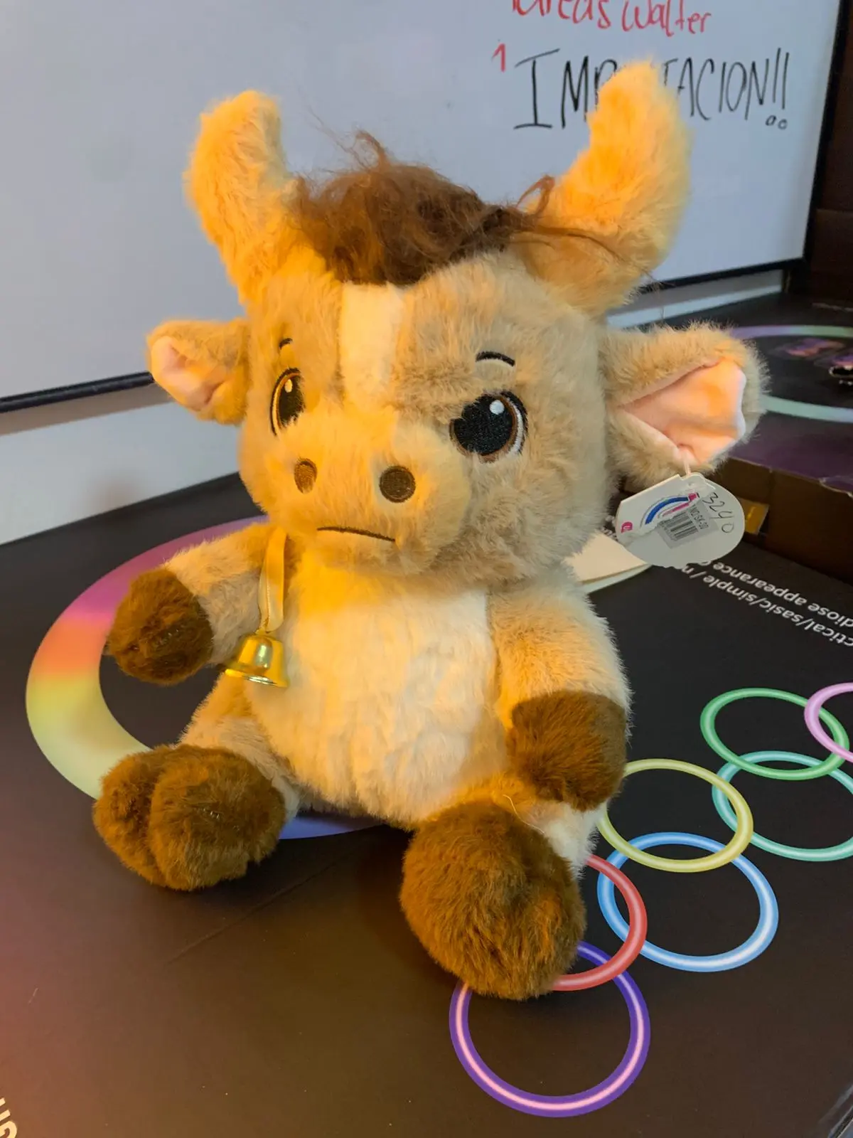 Peluche de toro beige con manchas marrones, cuernos suaves, copete desordenado, campana dorada al cuello y etiqueta colgante.