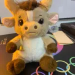 Peluche de toro beis y marrón con cuernos suaves, hocico detallado, campana dorada colgante y etiqueta en la oreja, sentado sobre superficie oscura.