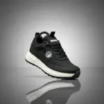 Zapatilla casual Brahma negra en material sintético con suela blanca y negra de PVC, vista lateral en ángulo flotando sobre superficie reflectante.