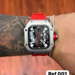 Reloj Richard Mille de aleación plateada y correa de silicona roja, con esfera negra esqueleto y detalles rojos, modelo Ref 001 en muñeca tatuada.