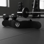 Arm Blaster negro para aislamiento de bíceps con logo de oso blanco, sobre piso de gimnasio con equipo de pesas al fondo.