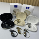 Audífonos inalámbricos ergonómicos en negro, dorado y blanco, con estuches de carga abiertos y cajas de empaque al fondo.