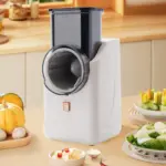 Picadora eléctrica de verduras blanca con tolva gris transparente, vista frontal en cocina con vegetales frescos y ensaladas picadas.