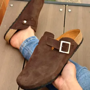 Zuecos casuales para hombre en cuero marrón ante, con hebilla lateral ajustable y suela corcho-caucho, mostrados en un pie sobre superficie de madera.
