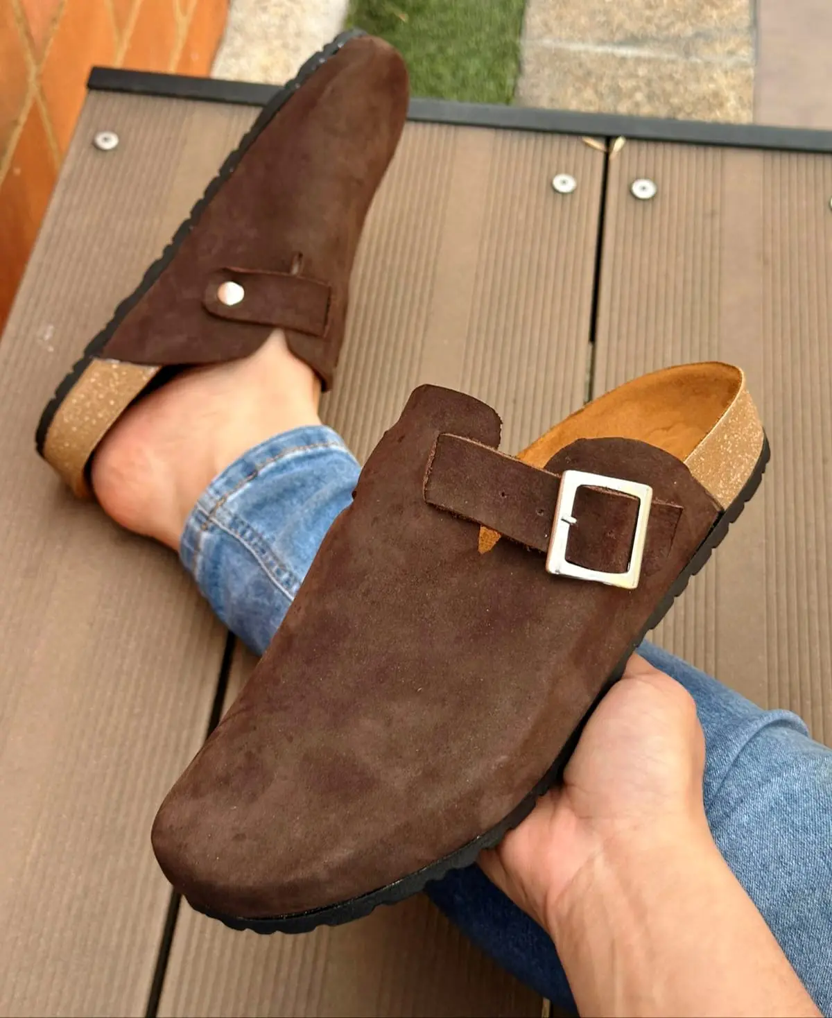 Zuecos casuales para hombre en cuero marrón ante, con hebilla lateral ajustable y suela corcho-caucho, mostrados en un pie sobre superficie de madera.