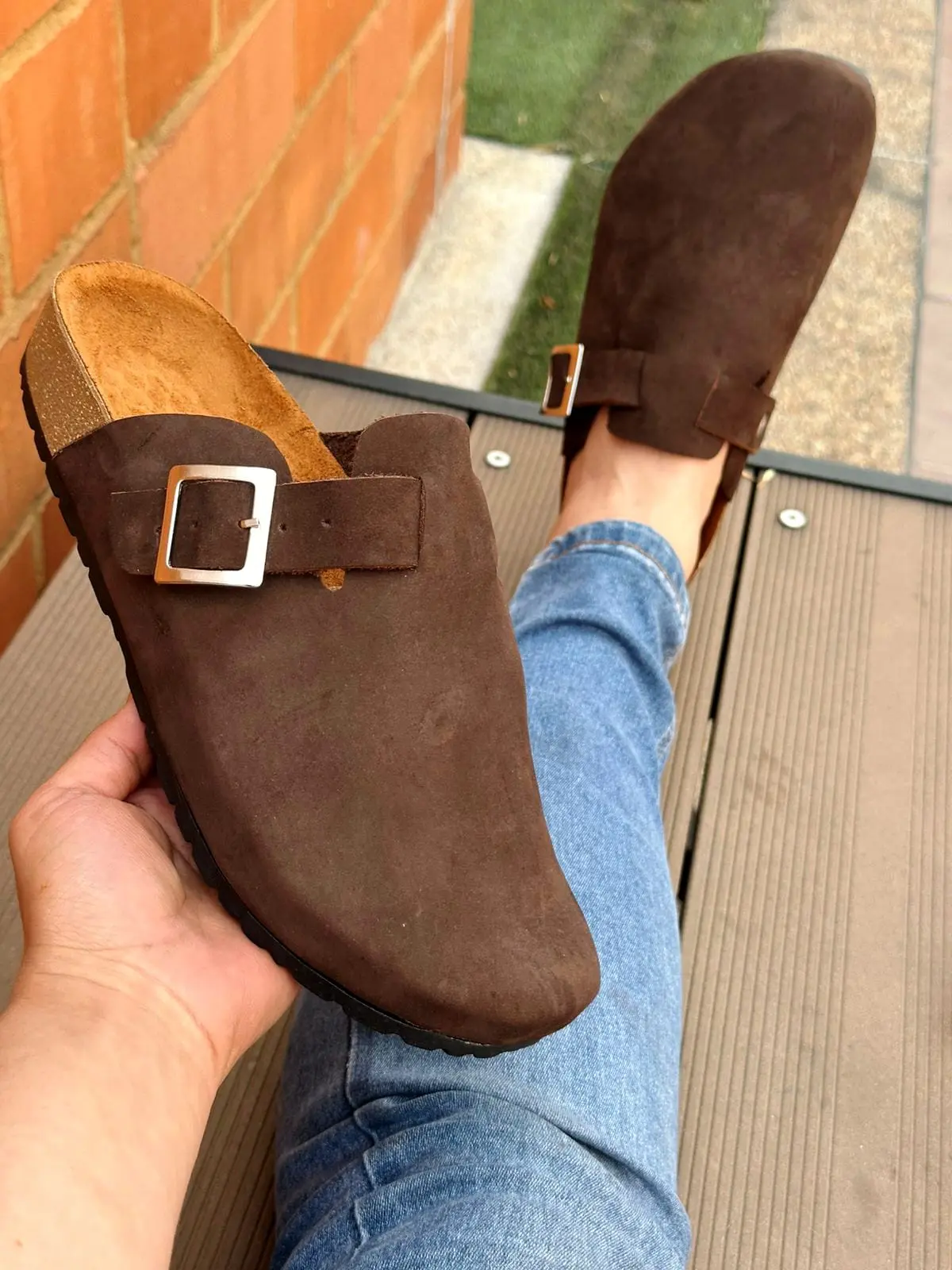 Sandalias suecos de cuero marrón oscuro tipo ante con hebilla plateada, sosteniendo una en la mano y otra puesta con jeans.