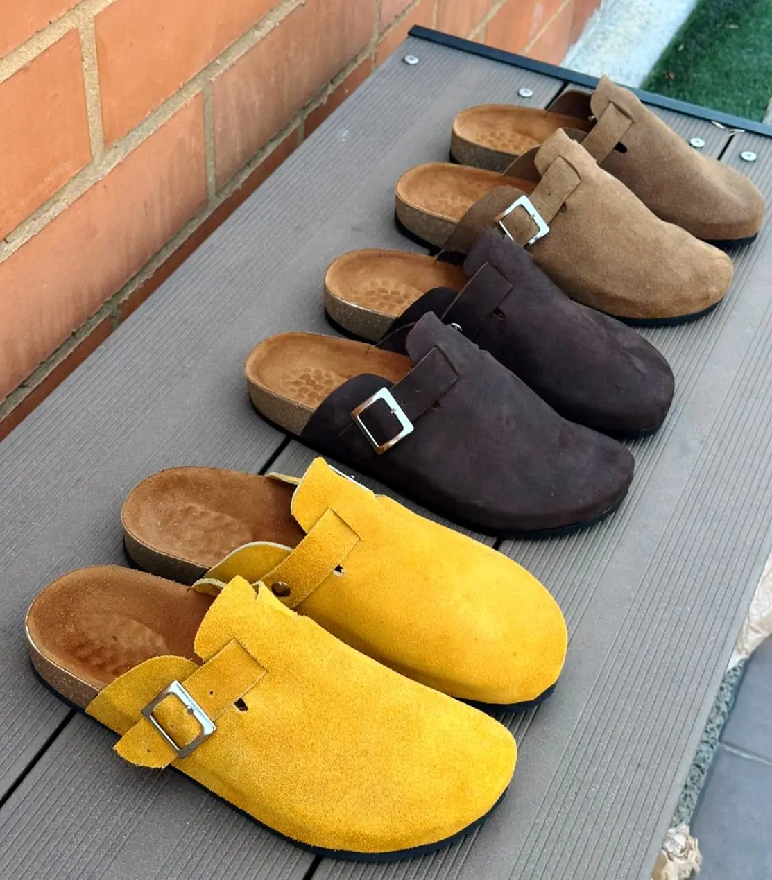 Sandalias suecos de cuero ante para hombre en colores amarillo mostaza, marrón oscuro y beige, dispuestas en diagonal sobre superficie de madera.
