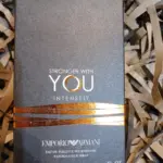 Caja gris oscuro con textura de Emporio Armani Stronger With You Intensely, acentos dorados y 100 ml sobre papel picado.