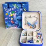 Set de manilla plateada con dijes coloridos en estuche rígido azul abierto, compartimentos con accesorios y bolsa de regalo temática de Stitch.