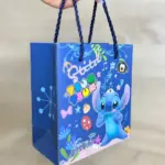 Estuche azul con diseño de Stitch y motivos coloridos, con asas de cuerda, sostenido por una mano sobre superficie de madera.