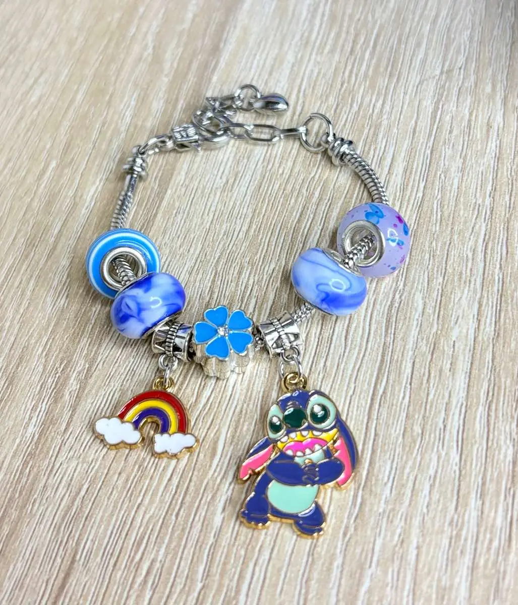 Manilla de cadena plateada con cuentas azules y blancas, dijes de flor azul, arcoíris y Stitch, sobre fondo de madera clara.