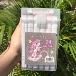 Estuche transparente con marcadores artísticos de doble punta y diseño de unicornio, sostenido frente a plantas.
