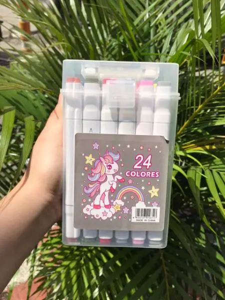 Estuche transparente con marcadores artísticos de doble punta y diseño de unicornio, sostenido frente a plantas.