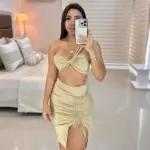 Conjunto beige de dos piezas con top bandeau cruzado de un hombro y falda ajustada con fruncido lateral y abertura en la pierna.