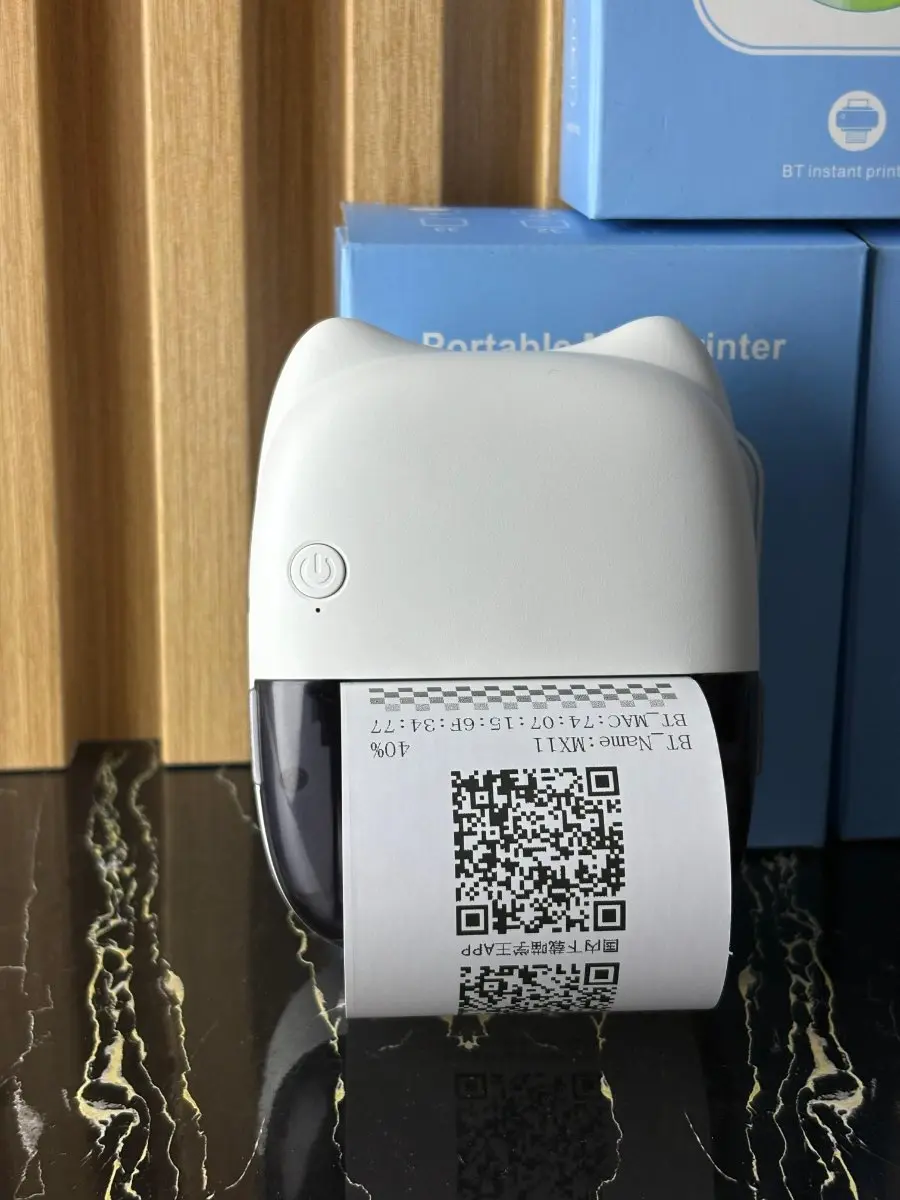 Mini impresora térmica portátil color blanco con diseño de orejas de gato, imprimiendo un ticket con código QR, sobre una superficie oscura.