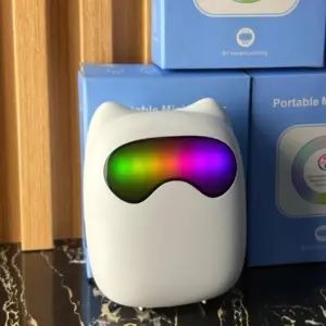 Mini impresora térmica Bluetooth portátil blanca con diseño de gato y luz RGB multicolor en la parte frontal, apoyada sobre superficie oscura frente a cajas azules.