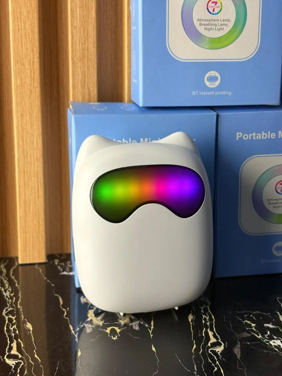 Mini impresora térmica Bluetooth portátil blanca con diseño de gato y luz RGB multicolor en la parte frontal, apoyada sobre superficie oscura frente a cajas azules.