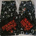 Dos pijamas de pareja negras con estampado de letras y luces navideñas, tela piel de durazno, con camisas de manga corta con el logo de Stranger Things en rojo.