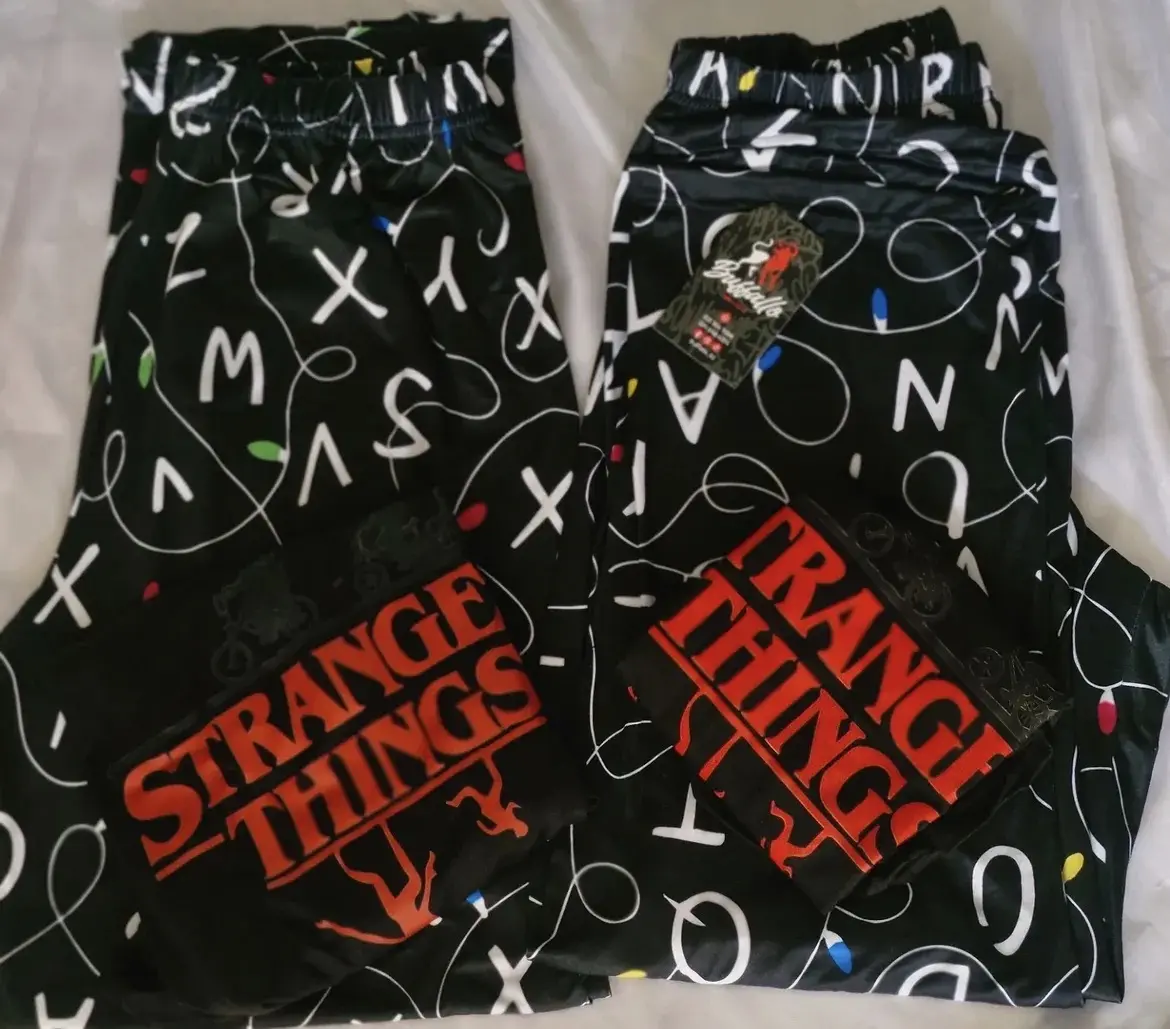 Dos pijamas de pareja negras con estampado de letras y luces navideñas, tela piel de durazno, con camisas de manga corta con el logo de Stranger Things en rojo.