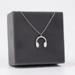 Collar de acero inoxidable plateado con dije de audífonos, presentado sobre una caja de exhibición gris oscuro.